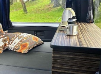 ハイエース車内のリビングスペース。収納ボックスを兼ねた木目調カウンターにケトルとマグカップ、窓から緑の景色を望む